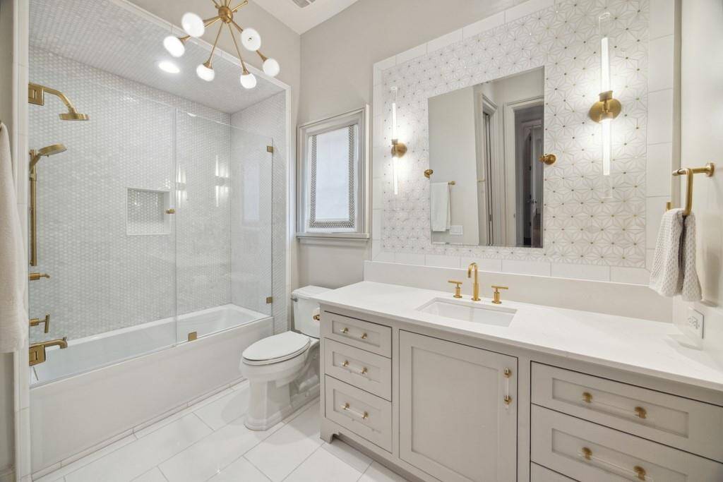 One of five full bathrooms, this beautifully designed space features gold hardware, designer lighting, a custom tile accent wall, and a frameless glass tub/shower with mosaic tile surround