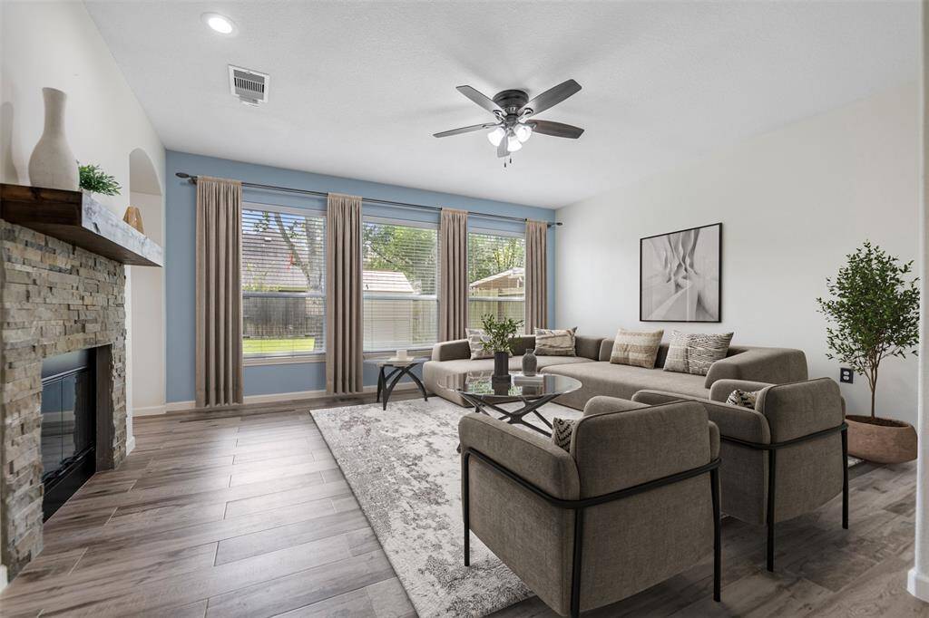 This spacious living room features three large windows that fill the space with natural light, wood-look flooring for a warm, modern touch, and a stunning stone fireplace with a classic mantle as the focal point. (This photo is virtually staged)