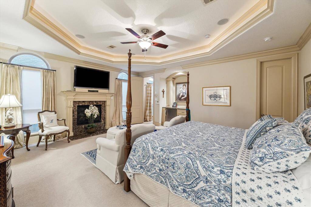 Open primary bedroom with trayed ceiling work, granite surround fireplace, custom fridge, and one of two closet spaces (closed door on the right). Fully carpeted for easy movement around.