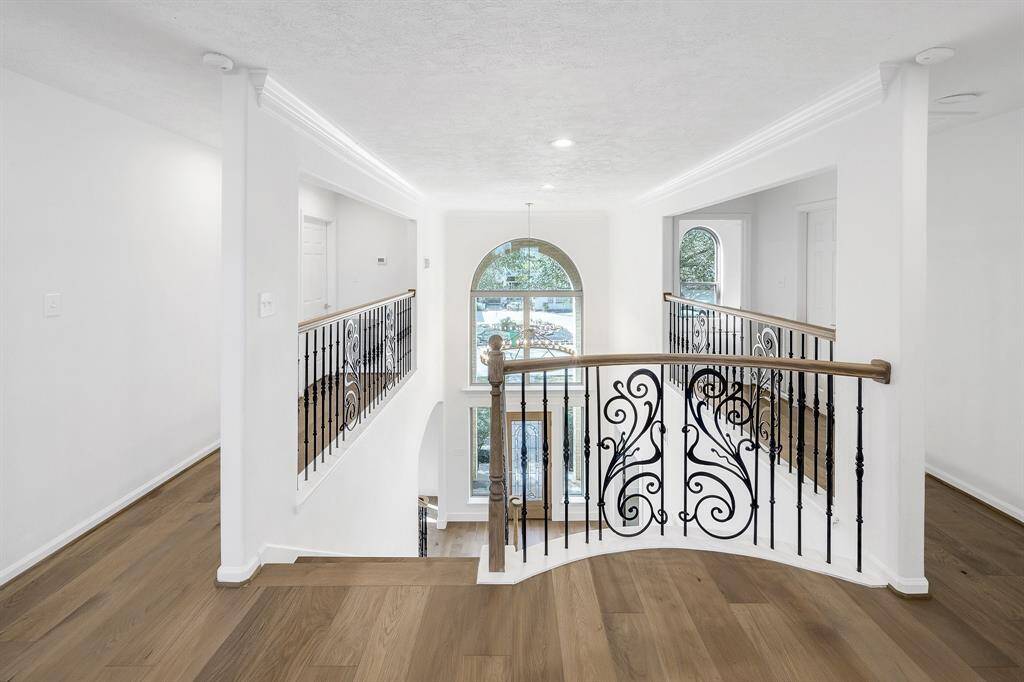 The upper hallway features gorgeous hardwood floors and intricate wrought iron railings with decorative scrollwork. Large windows and high ceilings fill the space with natural light, offering a perfect view of the grand entrance below.