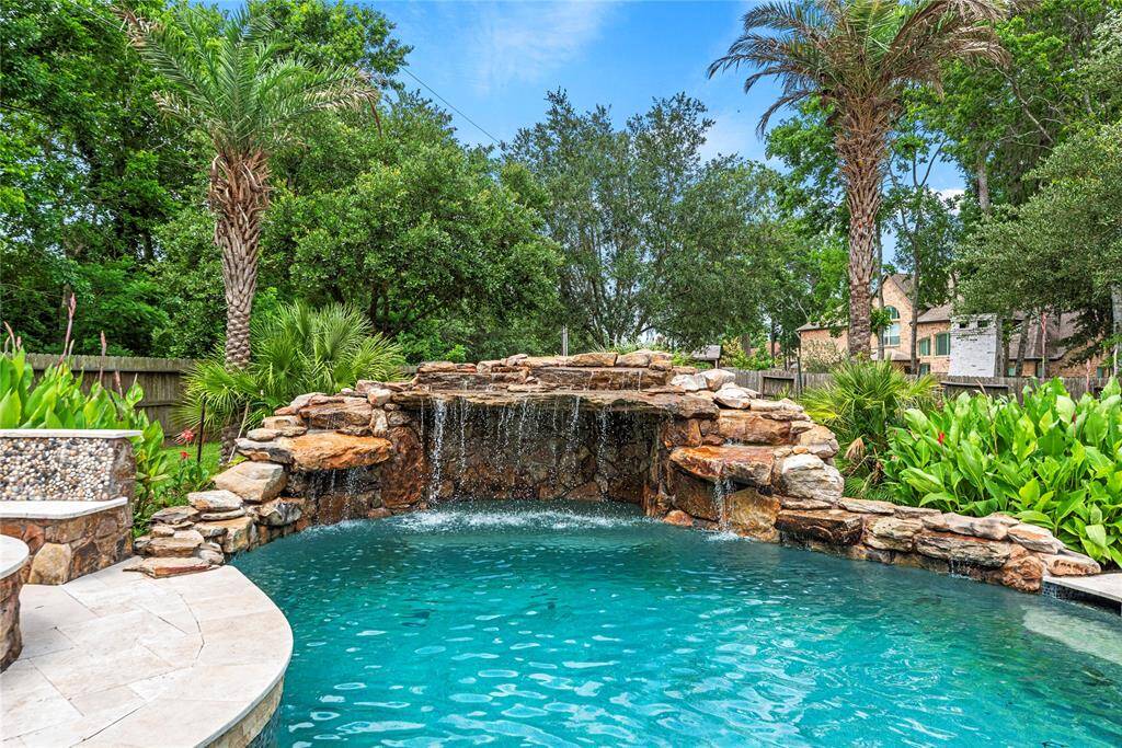 Rocks line the gently rounded edges of the sparkling swimming pool, now enhanced by a stunning grotto and waterfall feature. Reminiscent of a Bahamian retreat, vibrant green palm leaves complete the tropical paradise vibe.