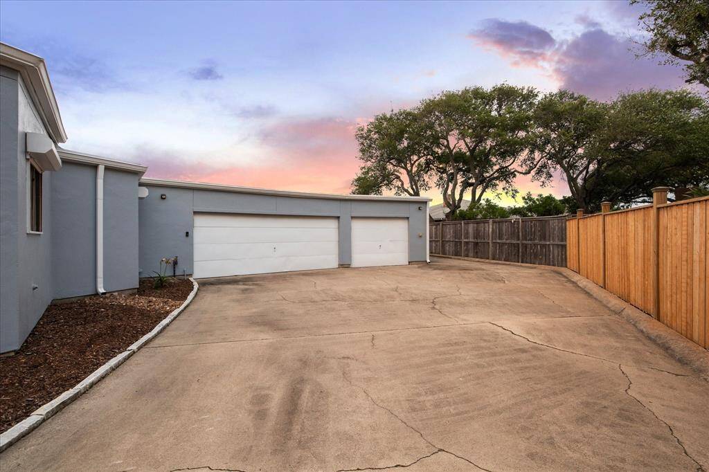 Spacious driveway leading to a three-car garage, surrounded by a wooden fence and mature trees, with a beautiful sunset sky in the background.