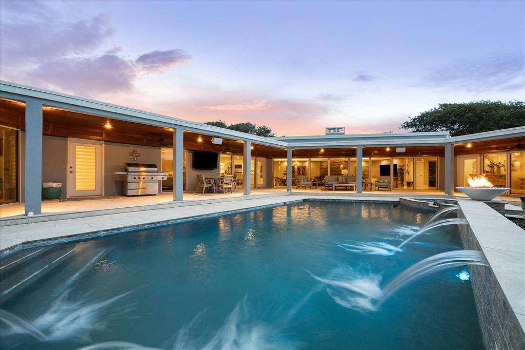 This modern home features a stunning outdoor area with a large pool, elegant water features, and a spacious patio. The covered seating area, complete with a grill, is perfect for entertaining, all under a beautiful twilight sky.