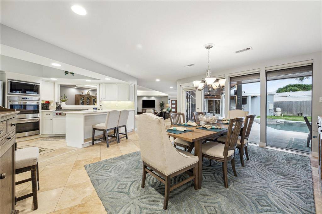 This open-concept space features a bright dining area with a large window view of a pool and backyard. The modern kitchen includes an island with seating and stainless steel appliances. The room is spacious and well-lit, ideal for entertaining.