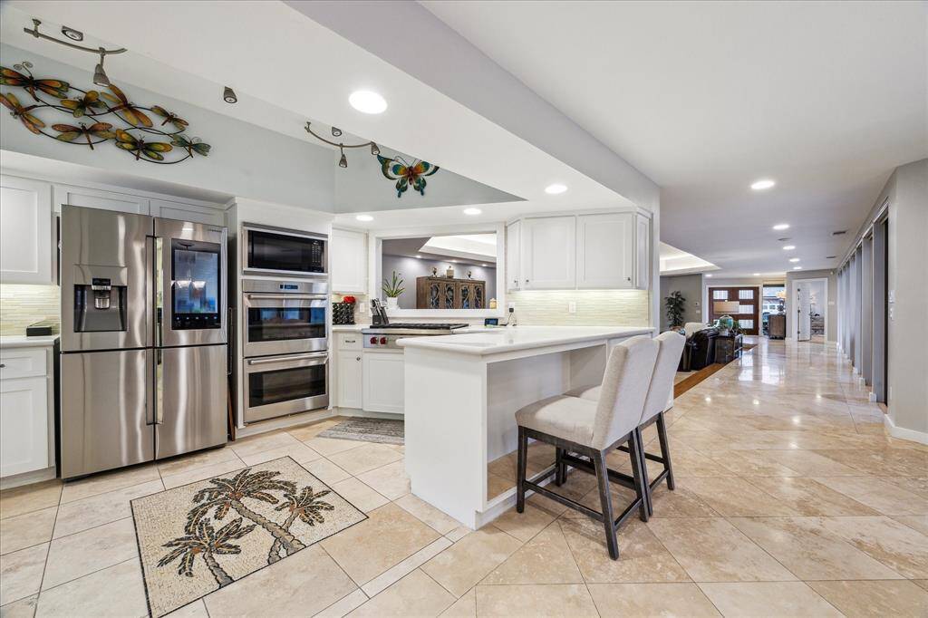 Modern kitchen with stainless high end appliances, white cabinetry, and a breakfast bar. Open layout leads to a spacious living area with tiled flooring, creating a bright and inviting space.