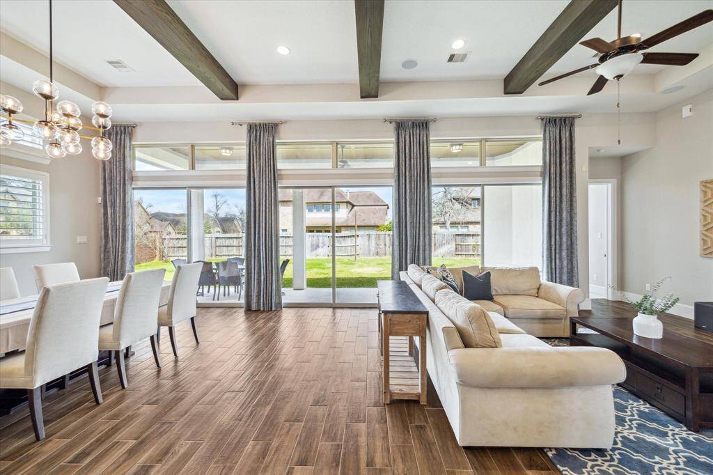 This stunning open-concept living and dining area features soaring 12' ceilings with rich wood beams, chic lighting, and elegant wood-look tile flooring. Expansive sliding glass doors disappear to seamlessly connect indoor and outdoor living, leading to the covered patio and backyard—perfect for entertaining or relaxing.