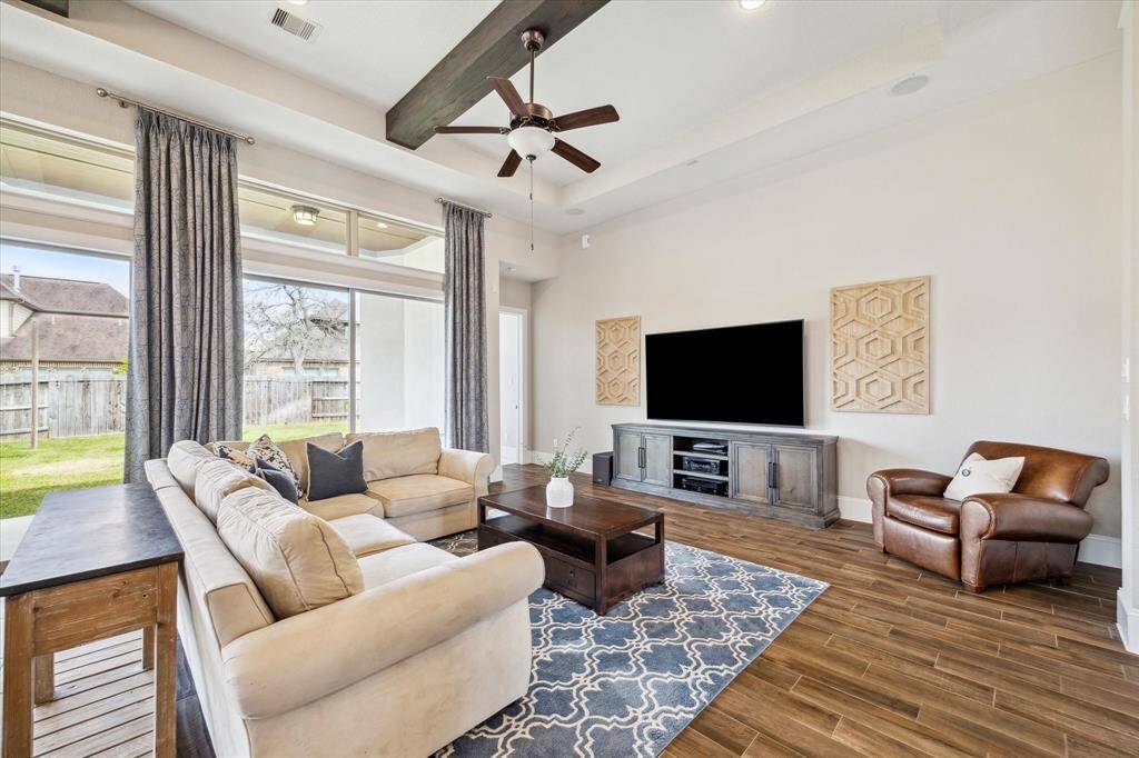 This inviting family room is a wonderful space to visit with friends and family or enjoy a quiet evening in. It's filled with neutral finishes including wood-look tile flooring, neutral paint, and a raised tray ceiling with rich wood beams, a modern fan, and upgraded lighting.
