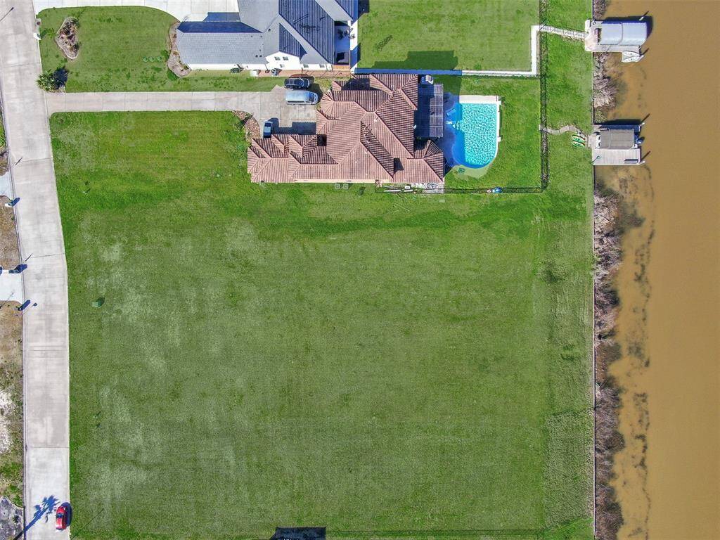 This aerial image shows a spacious, undeveloped lot adjacent to a residential property with a swimming pool. The lot is a large, open grassy area bordered by a street on one side and a water body on the other. It offers ample space for building a sizable home with potential for picturesque water views and a generous yard.
