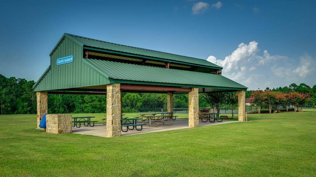 This is a spacious community pavilion with a green metal roof, stone pillars, and multiple picnic tables, set in a well-maintained park with lush green grass and trees in the background.
