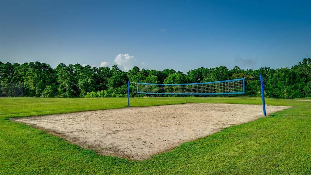 This photo features a well-maintained community volleyball court with a sand playing area and a blue net, set against a backdrop of lush greenery and trees under a blue sky. It suggests a family-friendly neighborhood with outdoor recreational amenities.