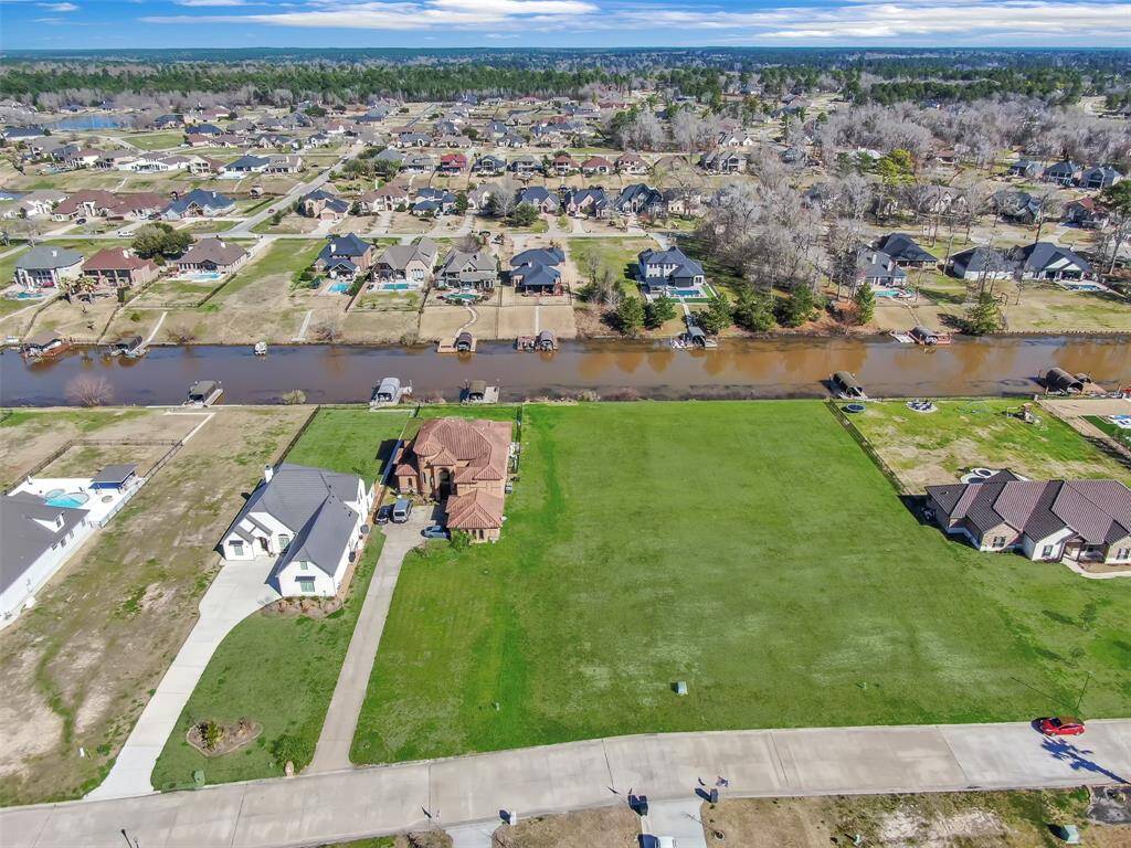 This aerial photo showcases a spacious, undeveloped green lot in a suburban neighborhood with existing homes nearby and a waterway adjacent to the property, offering potential for a custom-built home in a developed community.