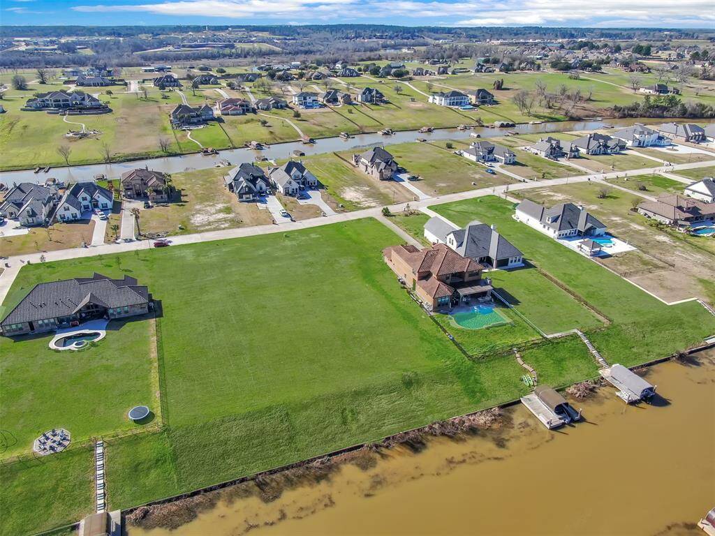 This aerial photo shows a spacious suburban neighborhood with large, detached single-family homes, expansive lawns, and a nearby waterway. Several homes feature backyard pools, and the community is laid out with wide streets and generous spacing between properties.