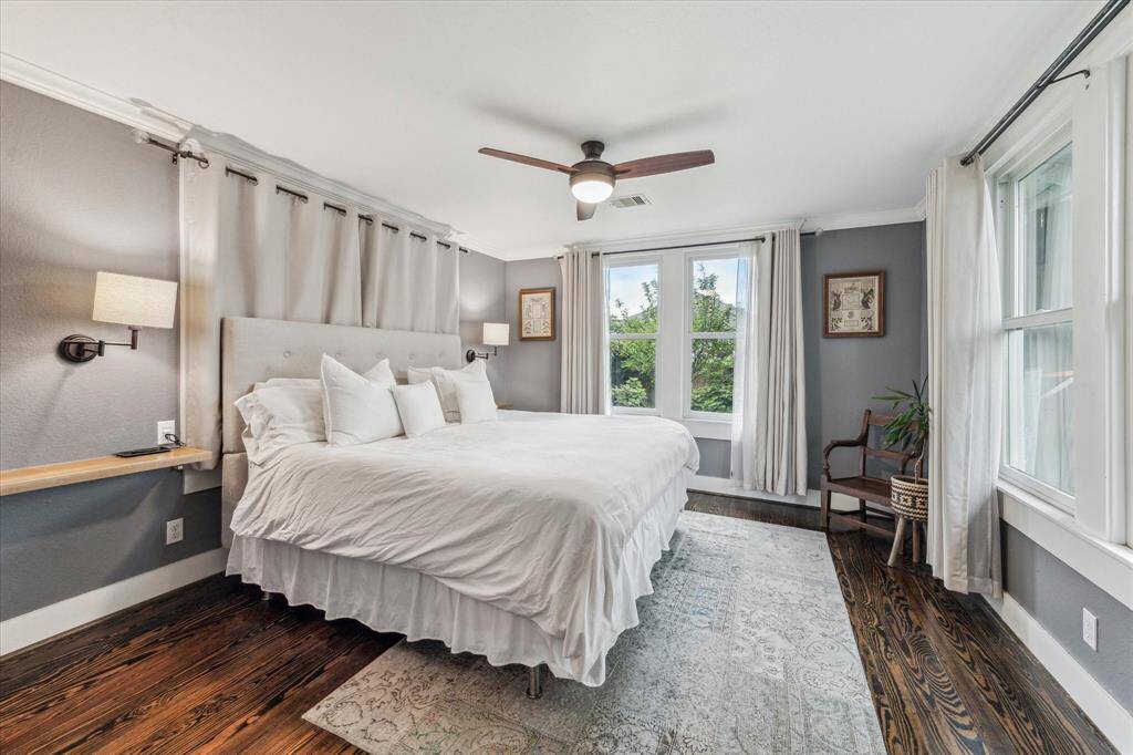 The spacious primary suite features hardwood floors, a ceiling fan, built-in reading lights, and serene views of the backyard and lush vegetable garden.