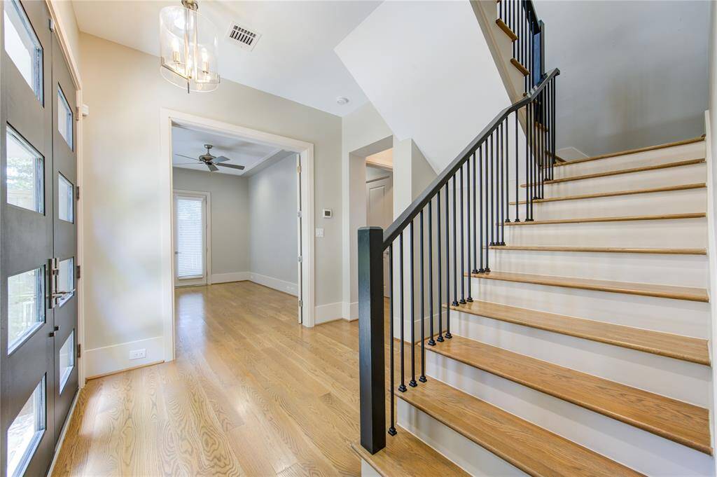 This photo showcases a bright and modern entryway with hardwood floors, a sleek staircase with black railing, and an adjacent room featuring a ceiling fan and large window.