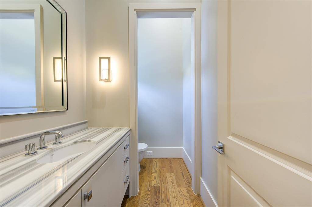 This is a modern bathroom with a sleek design featuring a marble countertop, a large mirror, and a wall-mounted light fixture. The wooden floor adds warmth, and the narrow layout leads to a separate toilet area.