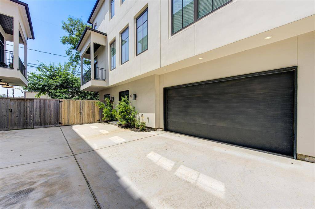 Modern townhouse exterior featuring a two-car garage with a sleek black door. The driveway is spacious, surrounded by a wooden fence and small landscaped area. Balconies and large windows enhance the clean, contemporary design.