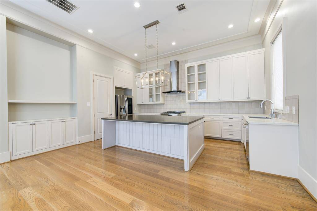 This spacious kitchen features hardwood floors, a large center island with a modern pendant light fixture, and ample white cabinetry. It includes stainless steel appliances, a built-in cooktop, and a sleek range hood. The room is bright and airy with recessed lighting and large windows.