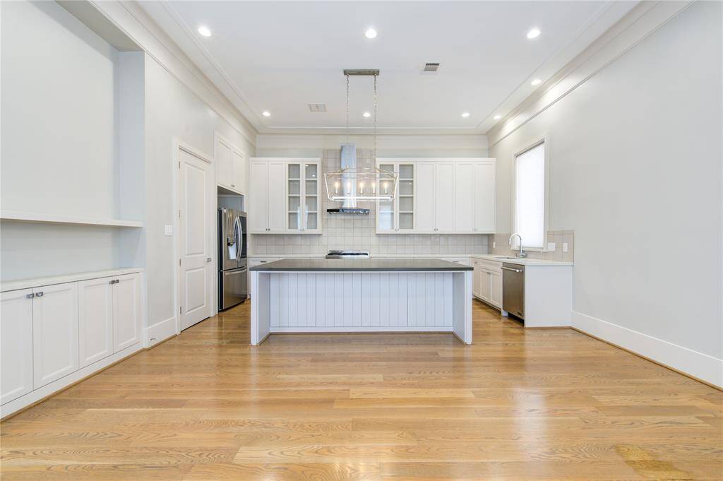 This spacious kitchen features hardwood floors, a large central island, modern stainless steel appliances, and ample white cabinetry. The room is bright with recessed lighting and a window for natural light.