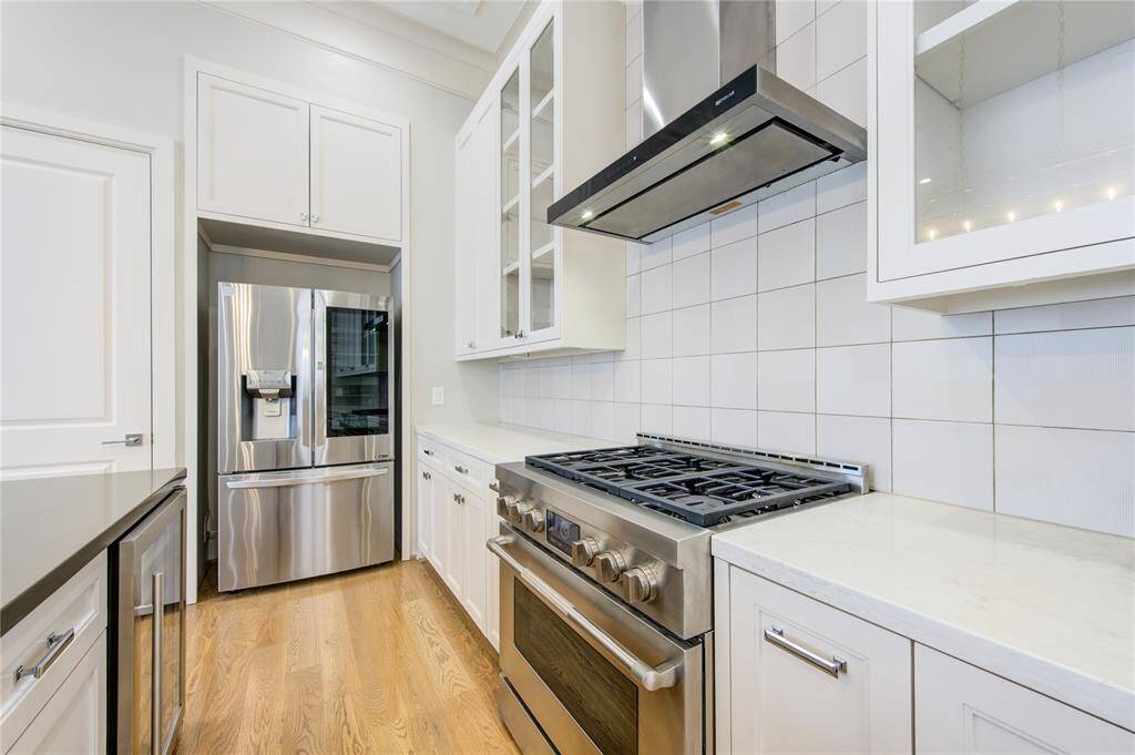 Modern kitchen with stainless steel appliances, including a large fridge and gas stove. Features sleek white cabinetry, tile backsplash, and hardwood floors, creating a clean and inviting space.
