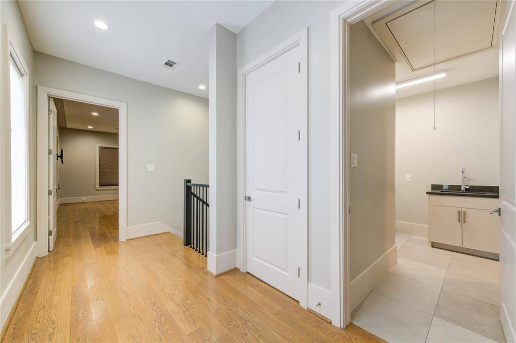 This photo shows a well-lit hallway with light wood flooring and neutral wall colors. It features a staircase with a black railing, a closed door, and an open door leading to a small room with a sink, suggesting a utility or laundry space. The hallway connects to another room with a window, adding to the home's spacious feel.