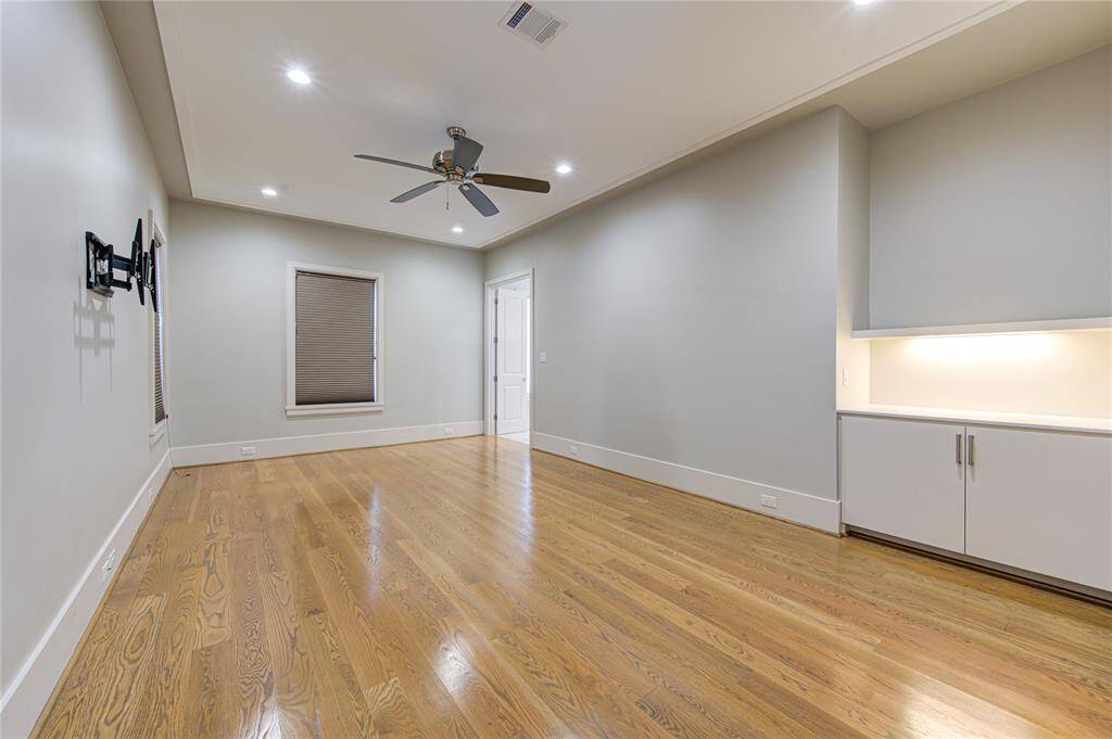 This room features hardwood floors, recessed lighting, and a ceiling fan. It includes a window with a blind, a wall-mounted TV bracket, and built-in cabinetry with under-cabinet lighting, offering a modern and functional space.