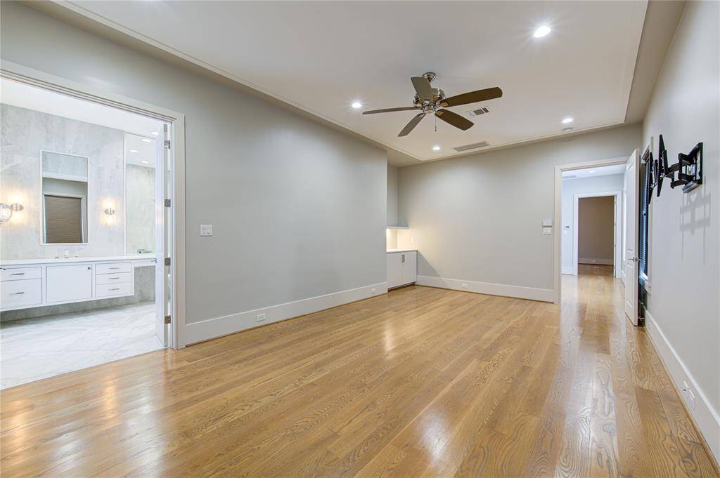 Spacious room with hardwood floors and neutral walls, featuring recessed lighting and a ceiling fan. It opens to a modern bathroom with a double vanity and sleek finishes. Adjacent hallway leads to additional rooms, providing an open and airy layout.