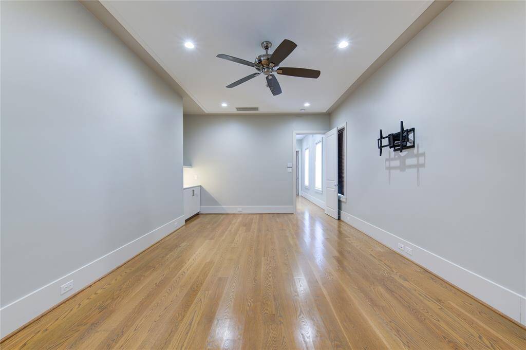 This room features hardwood flooring, recessed lighting, and a ceiling fan. It includes wall-mounted brackets for a TV and has neutral-colored walls, creating a clean, modern look.