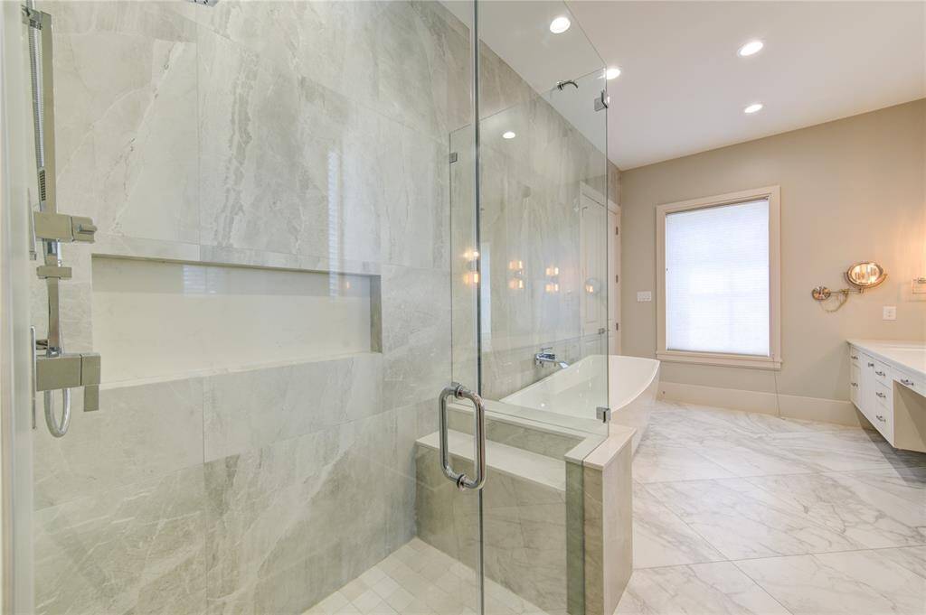 This modern bathroom features a spacious glass-enclosed shower with elegant marble walls, a freestanding bathtub, and a sleek vanity. Natural light streams in through a large window, highlighting the contemporary fixtures and bright, airy design.