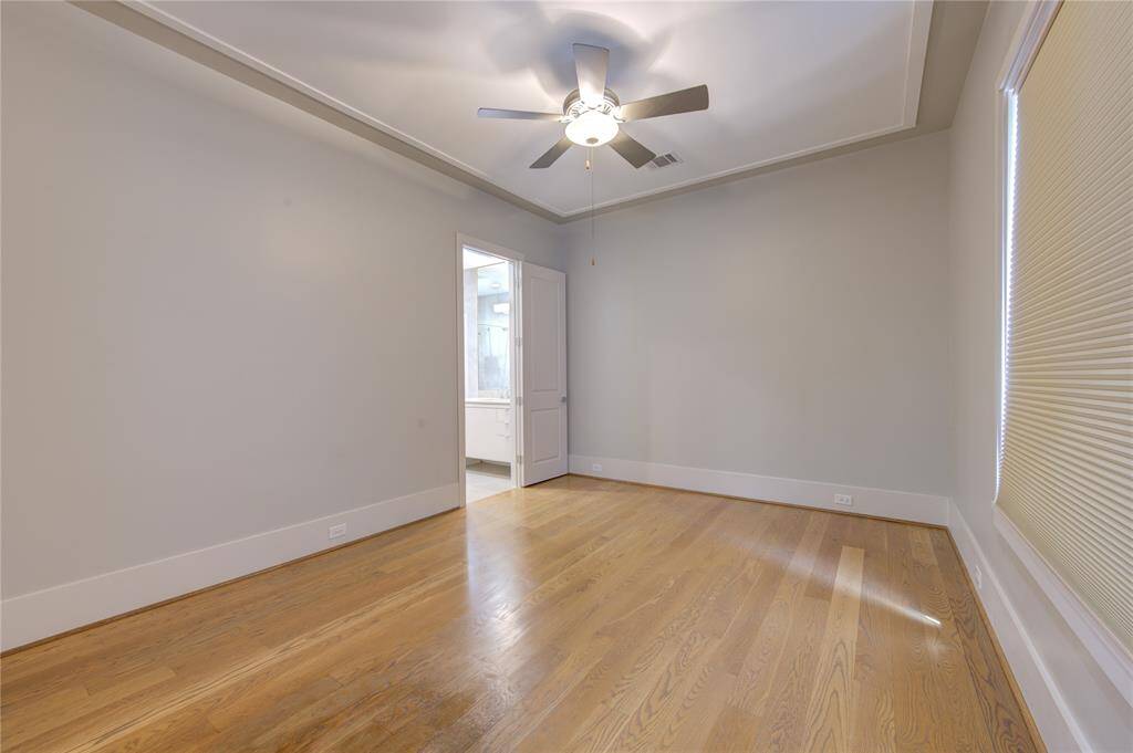 This photo shows a bright, spacious room with hardwood floors, neutral walls, and a ceiling fan. It features a large window with blinds and a doorway leading to an adjacent room, possibly a bathroom. Ideal for a bedroom or office space.