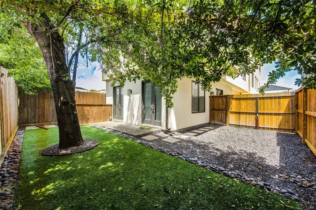 This photo showcases a charming backyard with artificial grass, a mature tree providing shade, and a gravel area. The space is enclosed by a wooden fence and features a modern exterior with large windows and patio doors, perfect for outdoor relaxation.