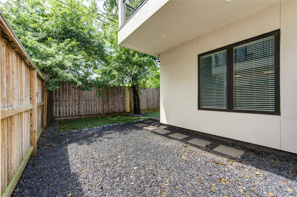This photo showcases a small, private backyard area with a gravel path and stepping stones, bordered by a wooden fence and shaded by trees. It features a modern exterior with large windows, providing a cozy outdoor retreat.