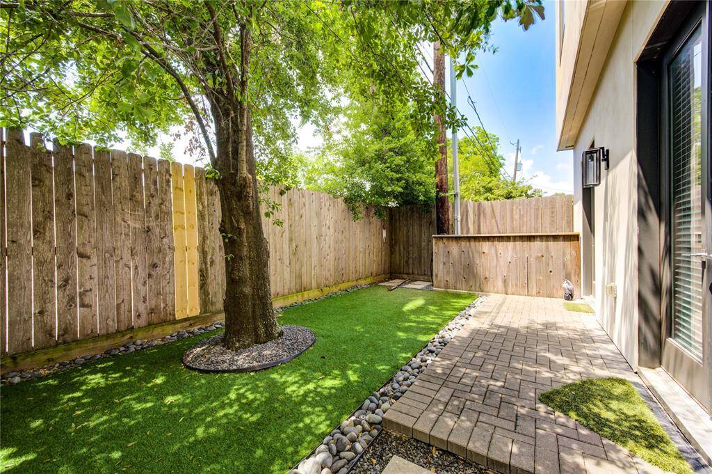 Charming backyard with a mix of artificial turf and a paved patio area, bordered by a wooden privacy fence. A mature tree provides shade, creating a serene outdoor space ideal for relaxation or entertaining.