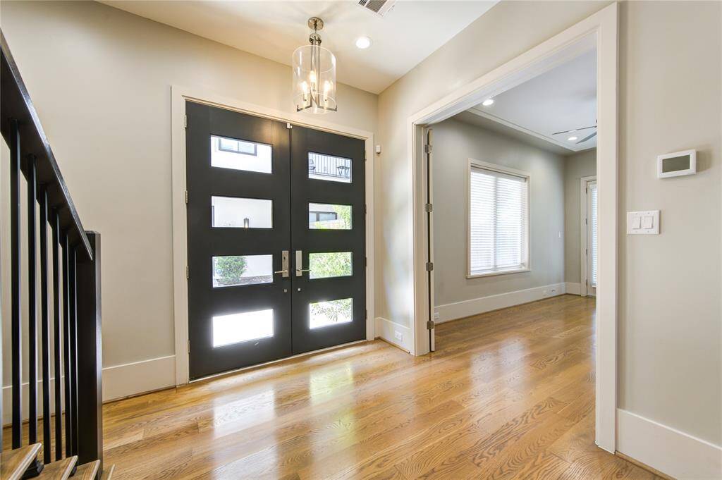 This entryway features a modern double door with glass inserts, providing natural light. It opens into a bright space with hardwood floors and neutral walls, leading to a room with a window and ceiling fan. A stylish light fixture and staircase add to the contemporary feel.