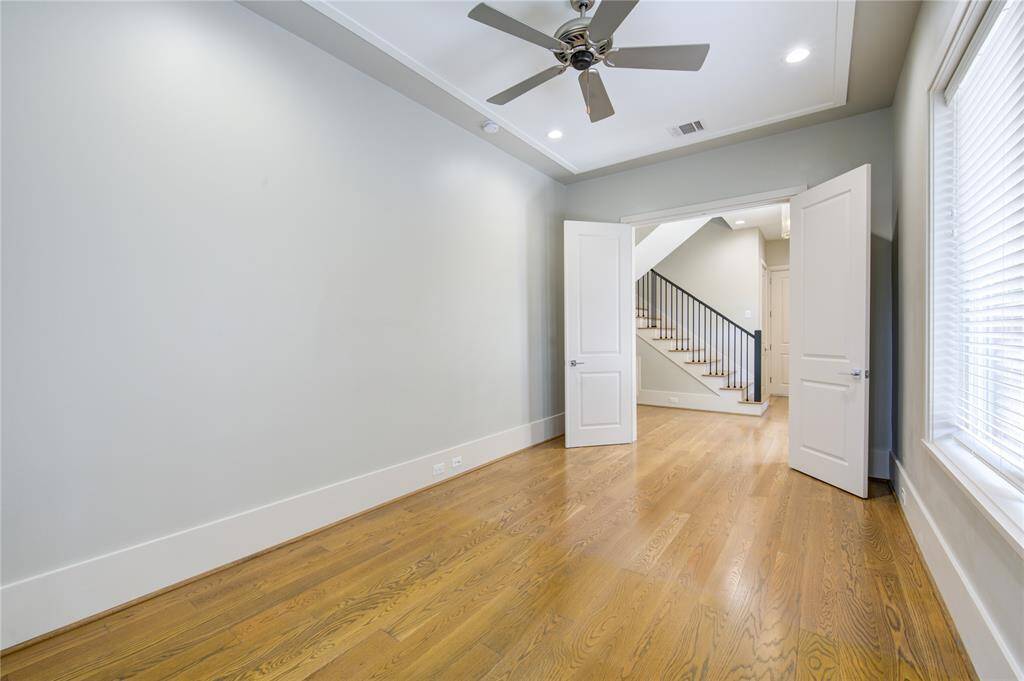 Bright, spacious room with hardwood floors, neutral walls, and a ceiling fan. Features large windows for natural light and double doors leading to a staircase with a modern railing.