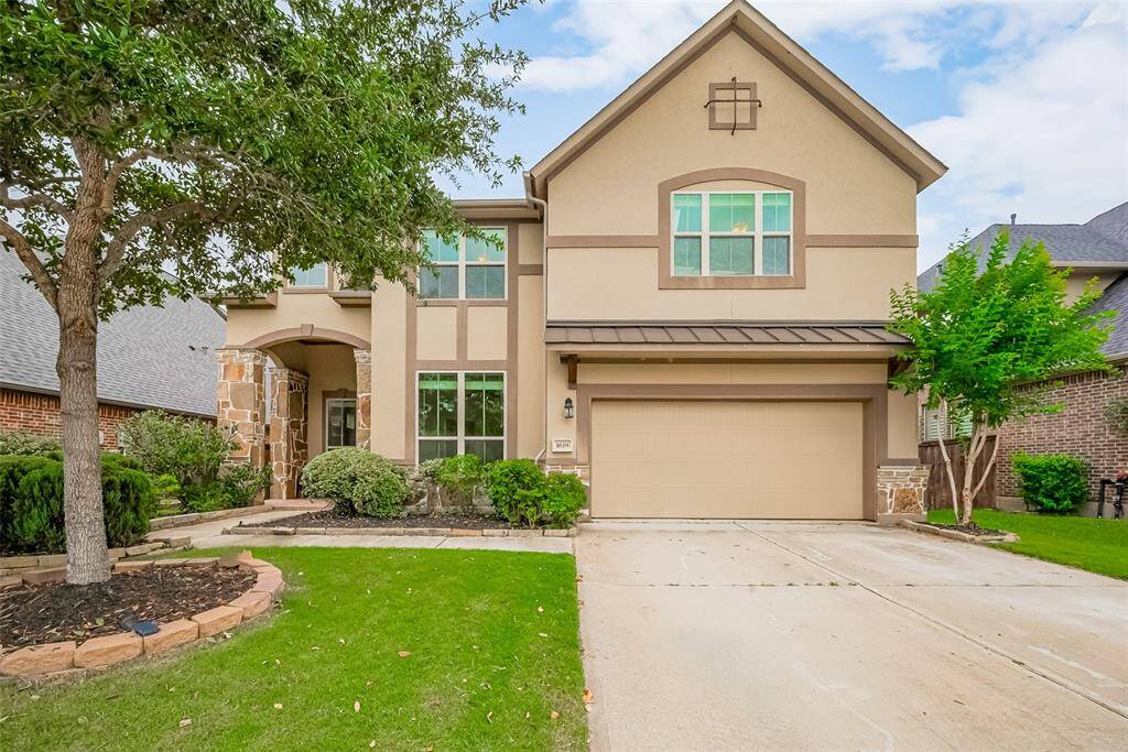 This is a two-story, modern home with a beige exterior and stone accents. It features a well-maintained front yard, a two-car garage, and a welcoming entrance with an arched doorway.