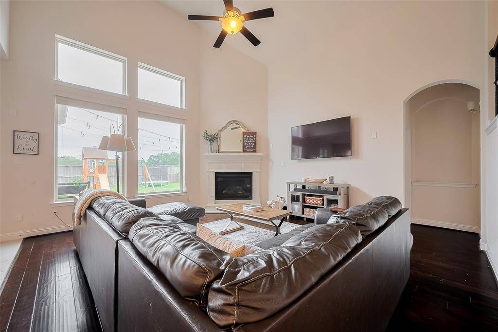 Bright and spacious living room with high ceilings, large windows, and a cozy fireplace. Features a modern ceiling fan, dark hardwood floors, and a view of the backyard playset. Ideal for family gatherings.