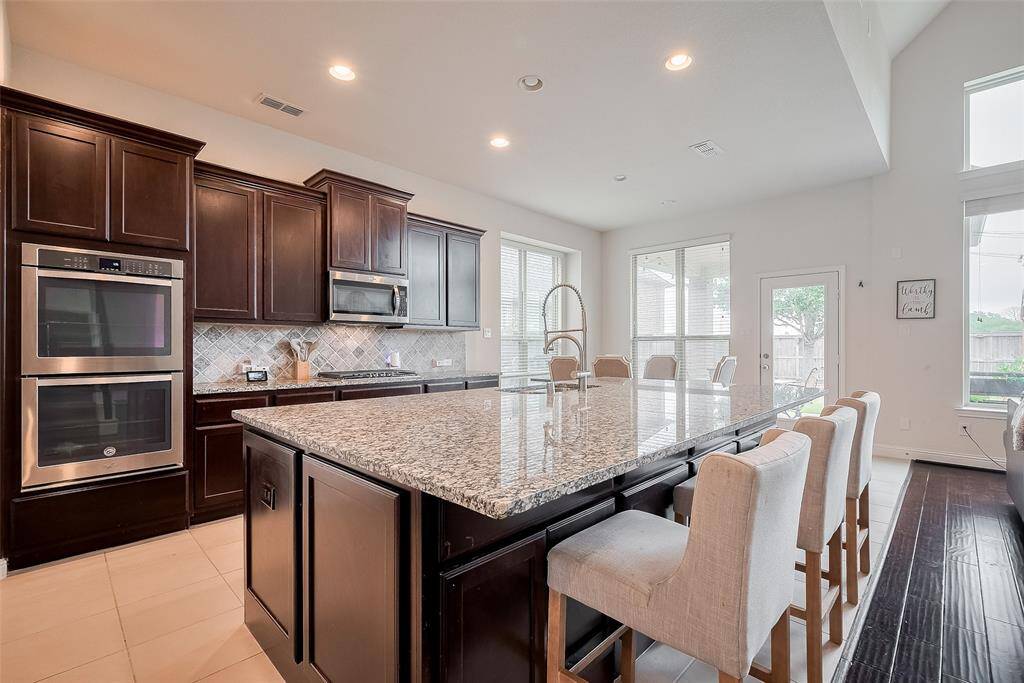 This kitchen features a spacious open layout with a large granite island, ideal for gatherings. It includes modern stainless steel appliances, dark wood cabinetry, and ample natural light from surrounding windows.