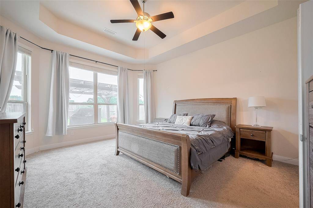 Bright and spacious bedroom with large windows, offering plenty of natural light. Features a tray ceiling with a fan, neutral carpeting.