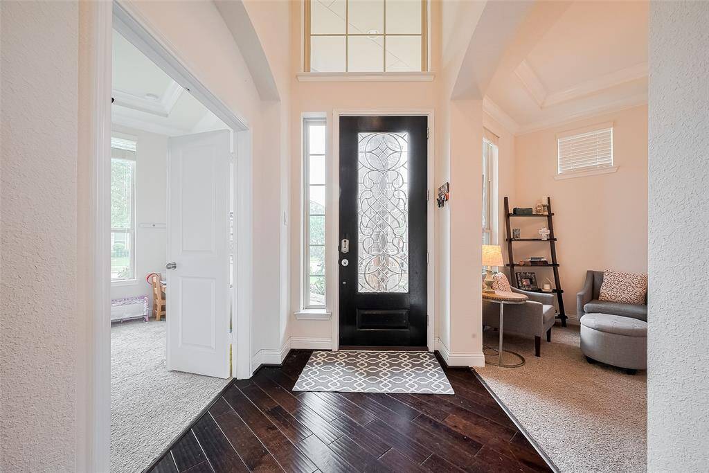This welcoming entryway features a stylish black front door with decorative glass panels, leading into a bright, open space with high ceilings. The area is complemented by rich dark wood flooring and adjacent cozy carpeted rooms, perfect for relaxation or a home office.