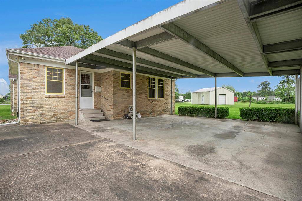This photo shows a brick home with a covered carport and a spacious driveway. The property features a neatly maintained lawn and a large, detached outbuilding in the background, offering ample outdoor storage or workspace.