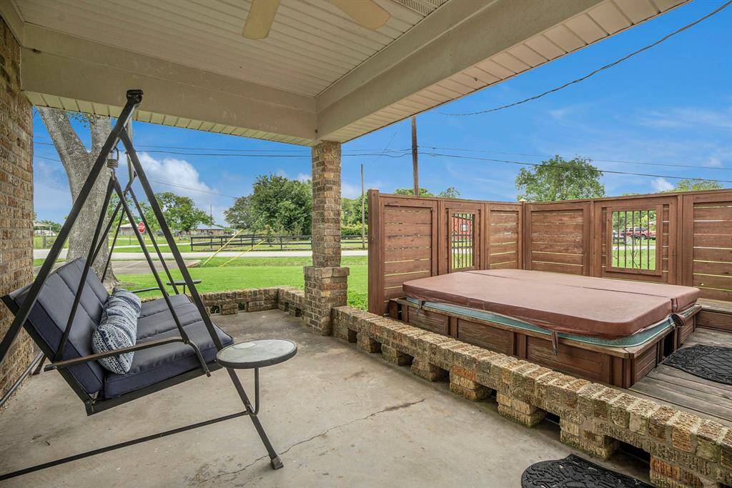 This charming outdoor space features a cozy porch with a hot tub, perfect for relaxation. It has a partially covered area with brick accents, a ceiling fan, and a wooden privacy fence, creating a welcoming and private retreat.