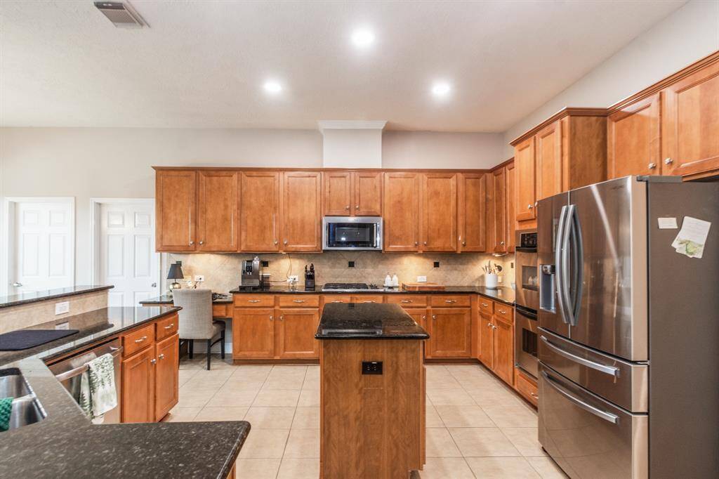 Spacious kitchen with warm wooden cabinets, granite countertops, and stainless steel appliances. Includes a central island and ample lighting, creating a welcoming and functional cooking space.