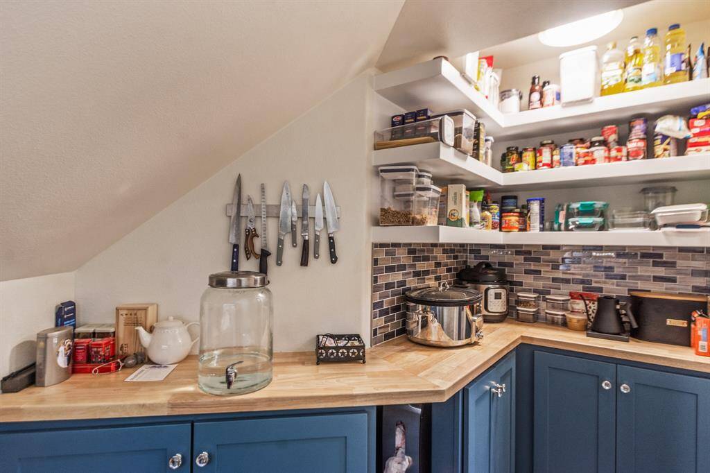 Step into a well-organized pantry with blue cabinets, butcher block countertops, and tiled backsplash. It features plenty of shelving, and kitchen essentials, including a knife rack and various appliances. The space is efficiently utilized, offering ample storage for culinary needs.