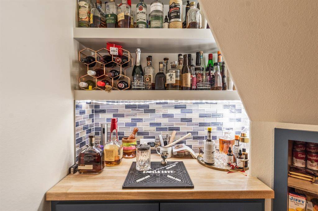 Cozy home bar tucked under a staircase, featuring a wooden countertop, stylish tile backsplash, and ample shelving for liquor and wine storage. Ideal for entertaining