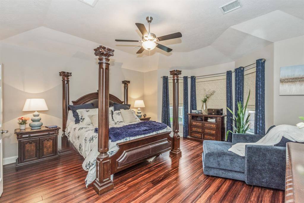 This spacious bedroom features elegant wood flooring, and ample natural light from multiple windows. It includes a cozy seating area, stylish furniture, and tasteful decor, creating a warm and inviting atmosphere.