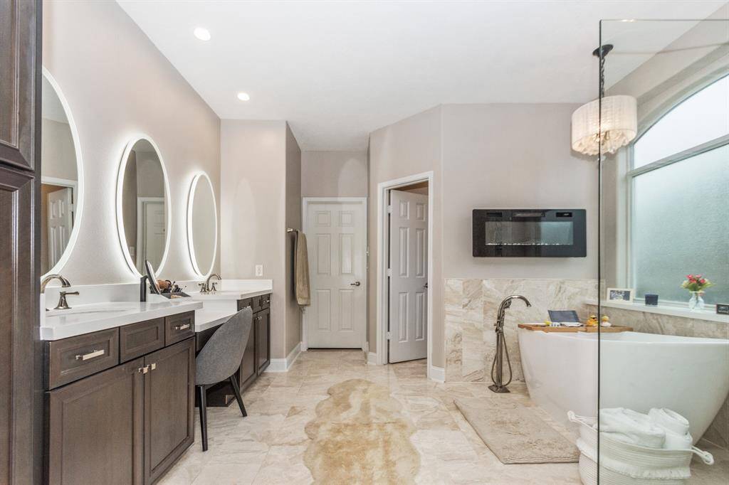 The master bathroom  features dark wood cabinetry with a double sink vanity and illuminated oval mirrors. The space is complemented by a neutral color scheme and marble flooring.