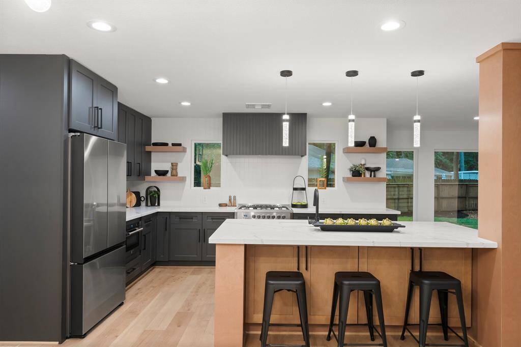 Well designed custom kitchen is anchored by central gas range with impressive vent hood, and symmetrical windows allowing natural light, natural wood shelving, and modern pendant lighting over large central island.