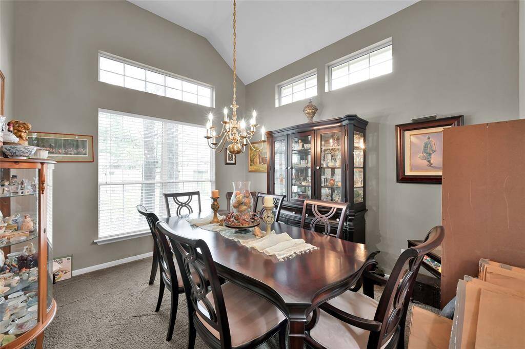 A bright, elegant dining room with high ceilings and large windows. It features a wooden dining table set, a chandelier, and display cabinets for decor. The room has a warm, inviting atmosphere with ample natural light.