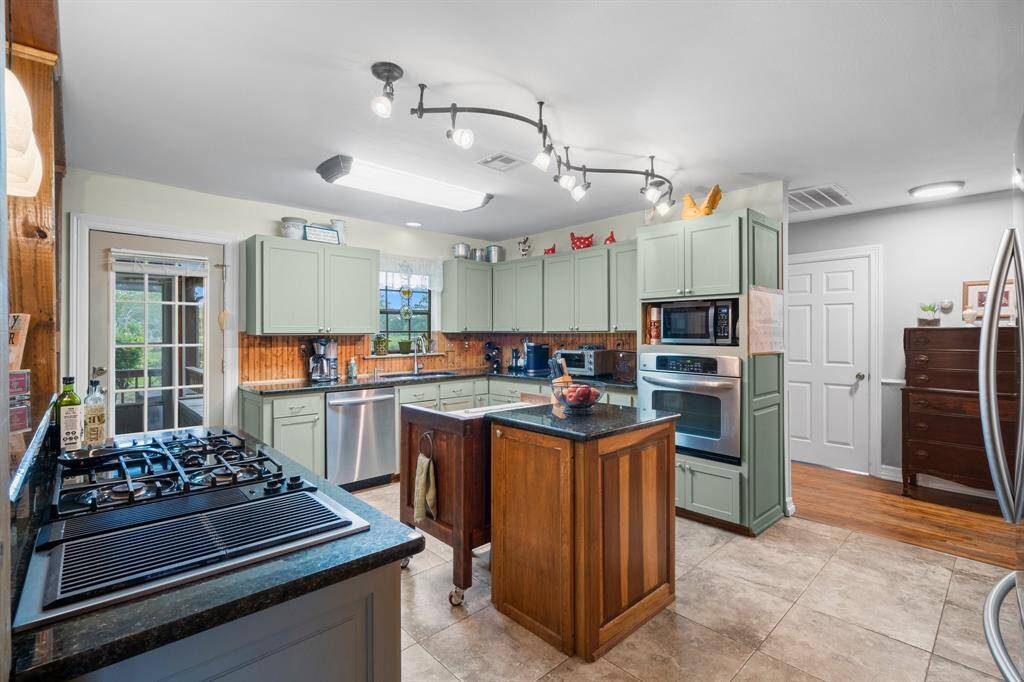 Charming kitchen with light green cabinets, stainless steel appliances, and a gas cooktop. Features include a central island, tile flooring, and ample natural light from a glass door leading outside. Perfect for both cooking and entertaining.