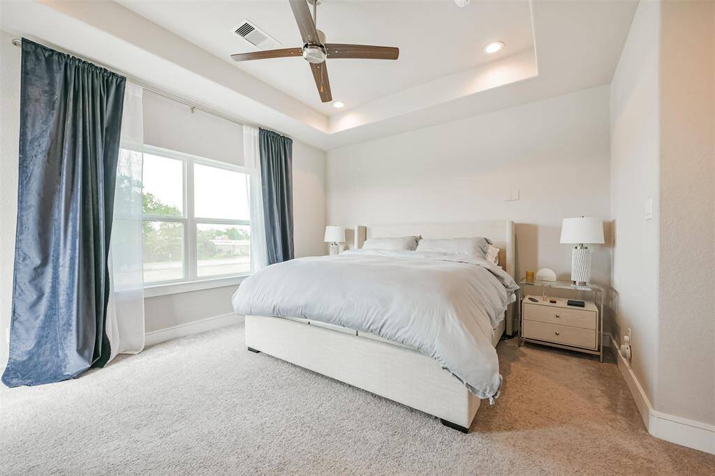 Bright and airy primary bedroom featuring plush carpeting, a large window with  for natural light, tray ceiling, a modern ceiling fan for comfort and five inch floor boards throughout for an added luxury feel.
